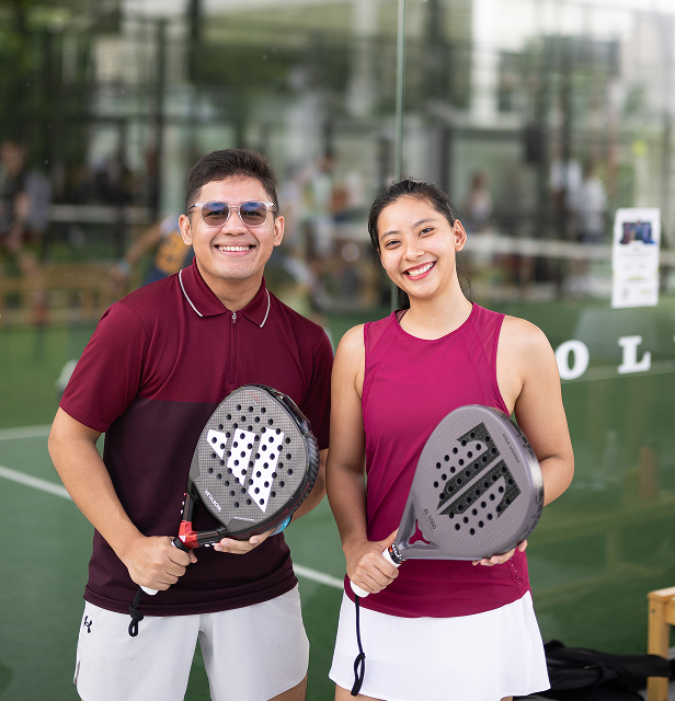 A man and a woman in matching maroon tops and white bottoms smile while holding padel rackets.